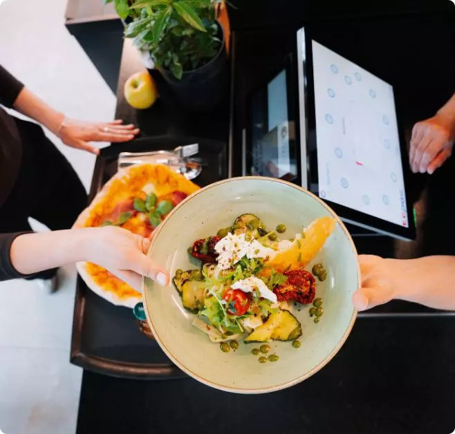 Bunte Salatschüssel mit frischem Gemüse, Käse und Kräutern wird über einer modernen Ausgabetheke serviert; daneben ist ein digitales Bestellterminal sichtbar.