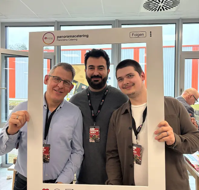Drei Personen posieren in einem hellen Veranstaltungsraum innerhalb eines großen, weißen Bilderrahmens im Stil eines Social-Media-Beitrags von „Panorama Catering“. Alle tragen Namensschilder und blicken in Richtung Kamera.