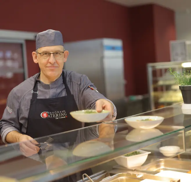 Person in Kochkleidung steht hinter einer Speisenausgabe und reicht eine Schale mit Speisen über die Theke. Auf der Schürze ist das Logo von Panorama Catering sichtbar.