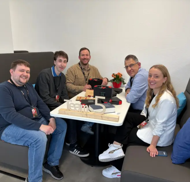 Fünf Personen sitzen an einem Tisch in einer Sitznische und blicken in Richtung Kamera. Auf dem Tisch stehen mehrere schwarze Boxen, Unterlagen und ein Holzaufsteller mit der Aufschrift „Team 5“.