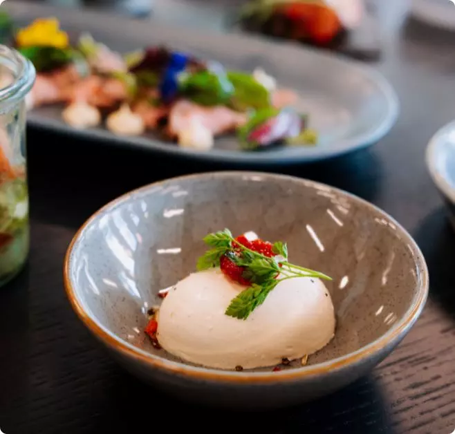 Modern angerichtetes Dessert in einer Keramikschale, garniert mit frischen Beeren und Kräutern. Im Hintergrund sind weitere hochwertige Speisen auf einem gedeckten Tisch zu sehen.